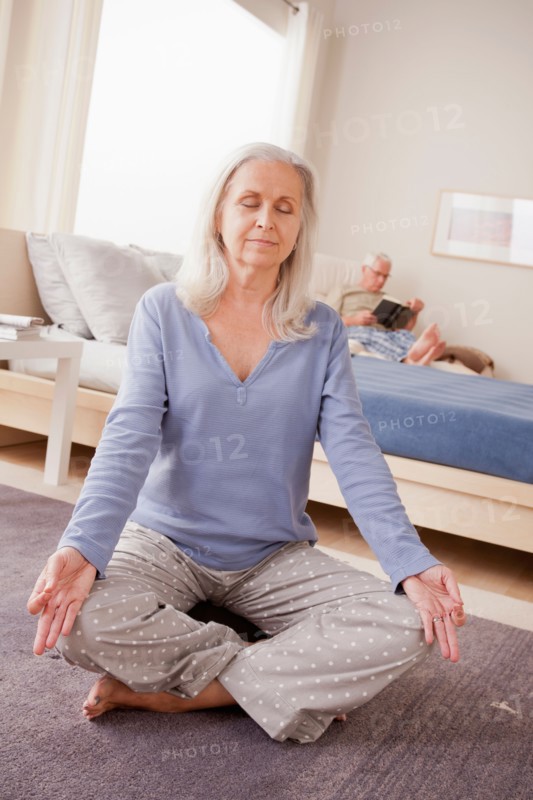 Senior woman practicing yoga with husband reading in background. Photo: Rob Lewine