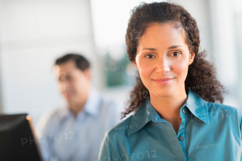 Portrait of businesswoman in office with male executive working in background.