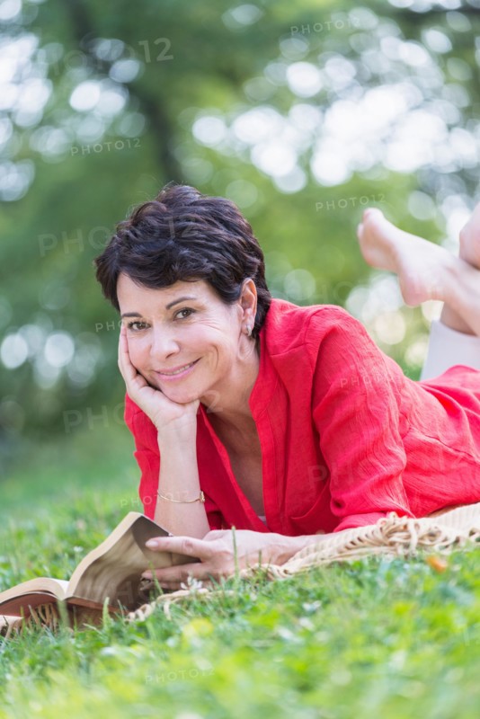 Mature woman lying on grass and reading book.