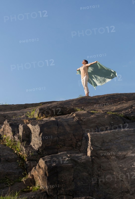 Mature woman standing on cliff with arms raised.