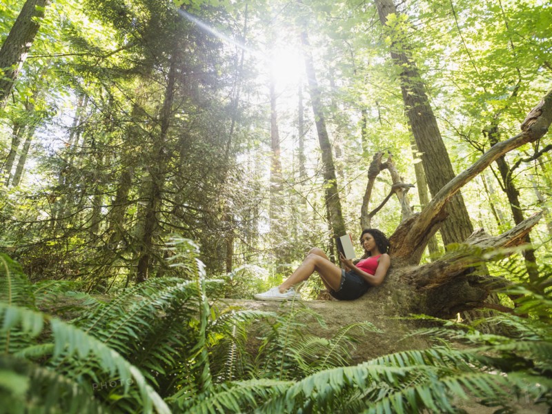 Young woman reading book on log in forest. USA, Oregon, Portland.