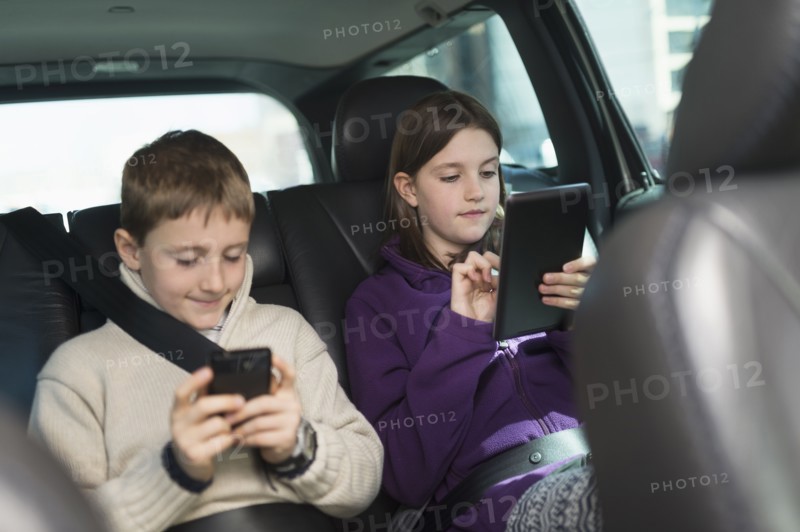 Boy and girl (8-9, 10-11) using digital tablet and smart phone in car.