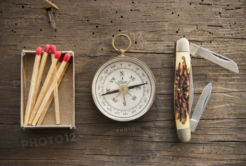 Studio Shot of matchsticks, compass and knife on wooden board.
Photo : Jamie Grill