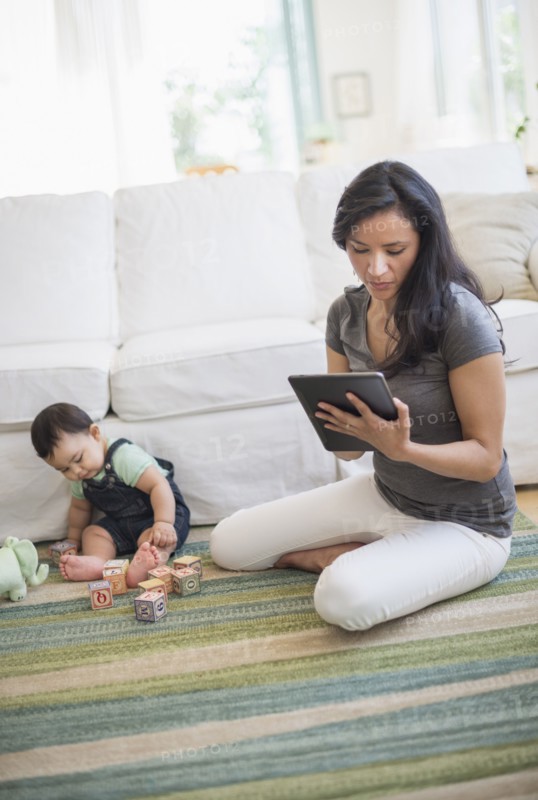 Mother using tablet pc while her son (6-11 months) playing with building blocks.