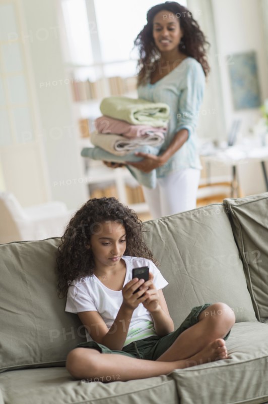 Girl (8-9) text messaging on sofa, mother in background.