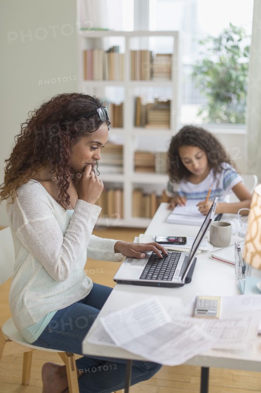 Woman using laptop at home, girl (8-9) doing homework in background.