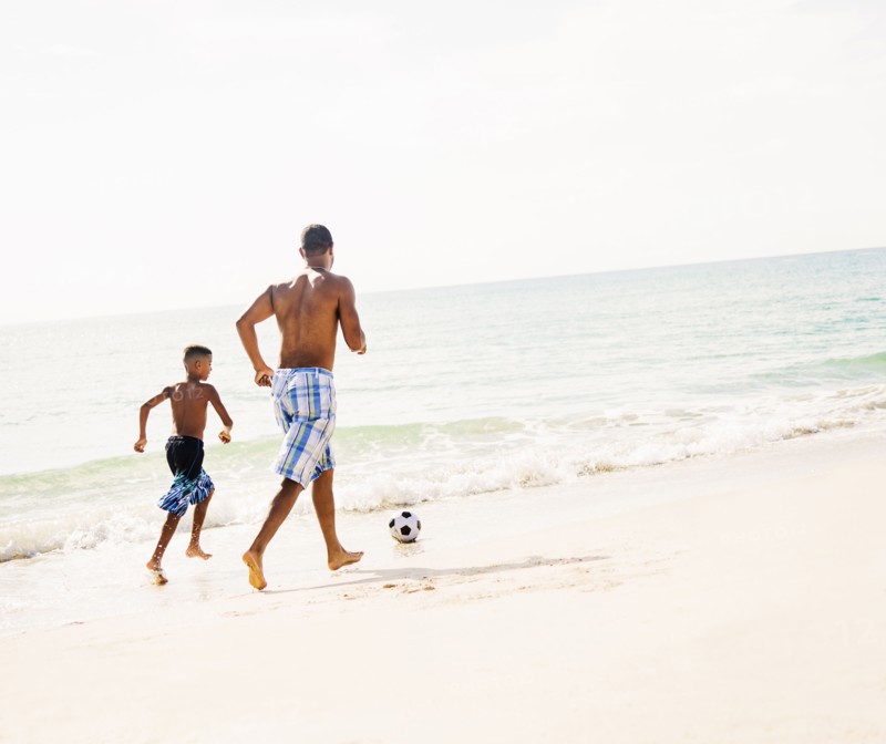Father and son (10-11) playing soccer on beach