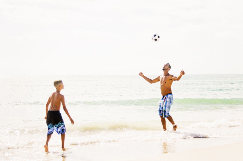 Father and son (10-11) playing soccer on beach