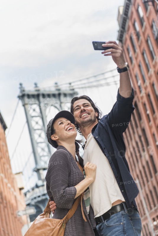 Couple taking selfie on street, Brooklyn Bridge in background. Brooklyn, New York.