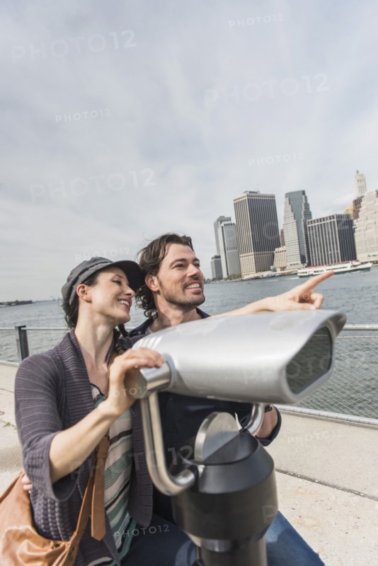 Happy couple sitting by coin-operated binoculars. Brooklyn, New York.