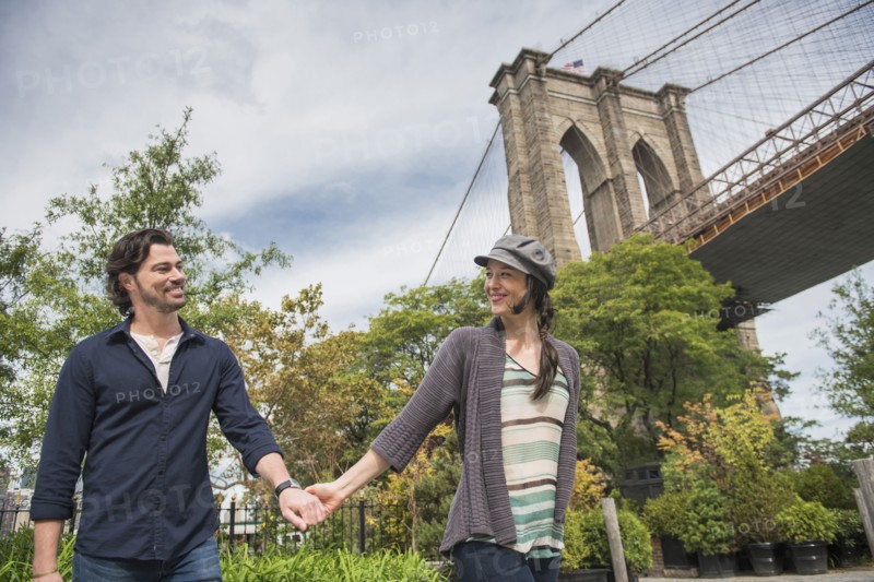 Happy couple holding hands and walking with Brooklyn Bridge in background. Brooklyn, New York.