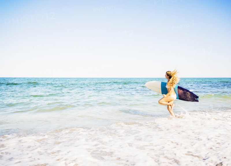 Young woman running in surf into sea carrying surfboard