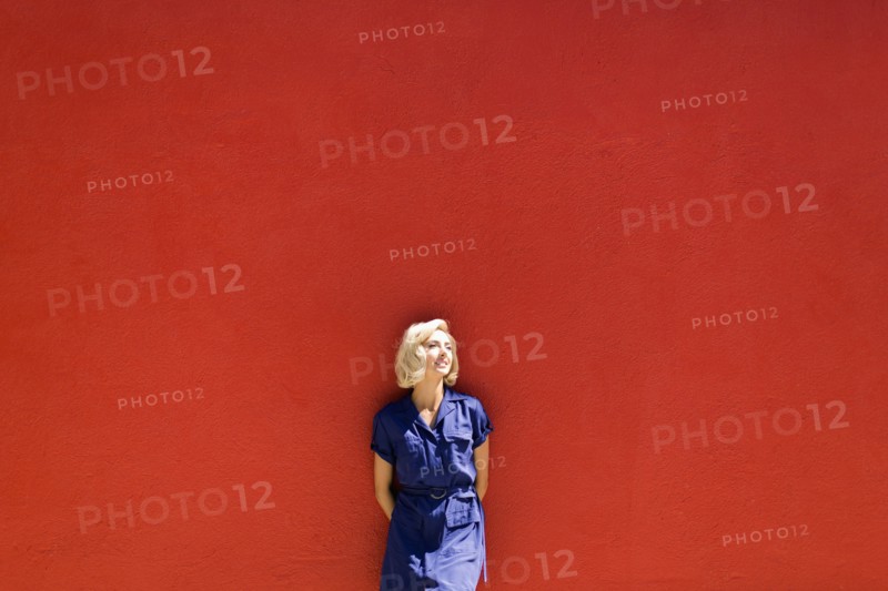 Woman in blue dress posing against red wall in sunlight