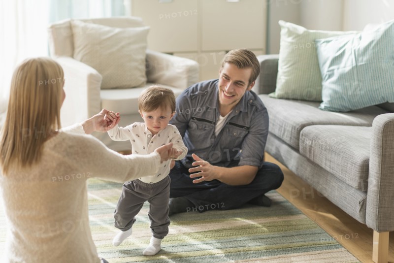 Happy parents helping little son (2-3 years) walking in living room.
