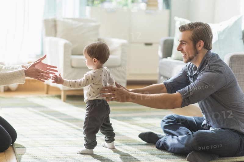 Happy parents helping little son (2-3 years) walking in living room.