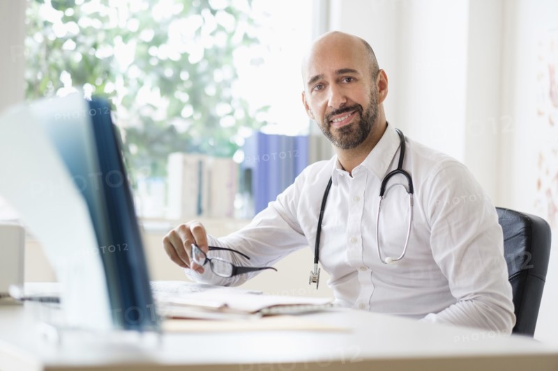 Portrait of smiley doctor working with laptop at desk in office.