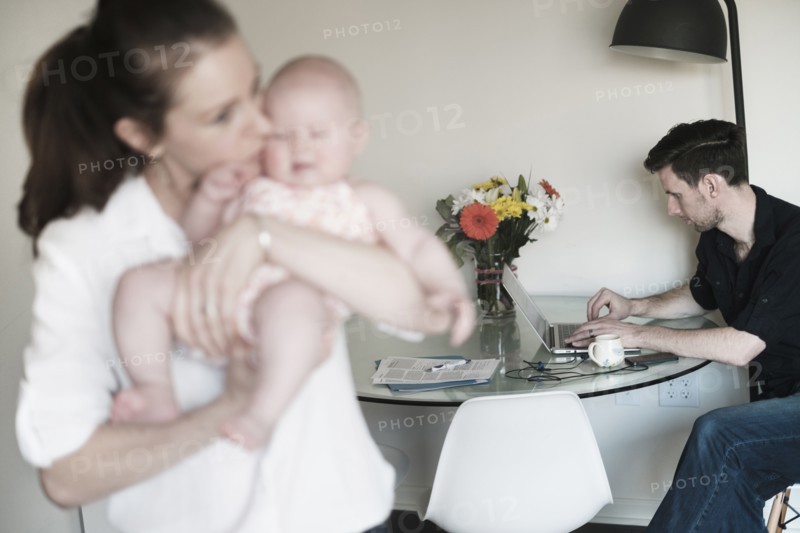 Mother holding baby (2-5 months) and father working in background.