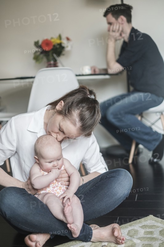 Mother holding baby (2-5 months) and father working in background.