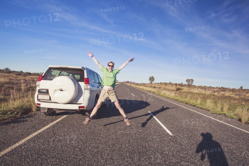 Australia, Outback, Northern Territory, Red Centre, Uluru-Kata Tjuta National Park, Woman jumping next to SUV car on road in wilderness