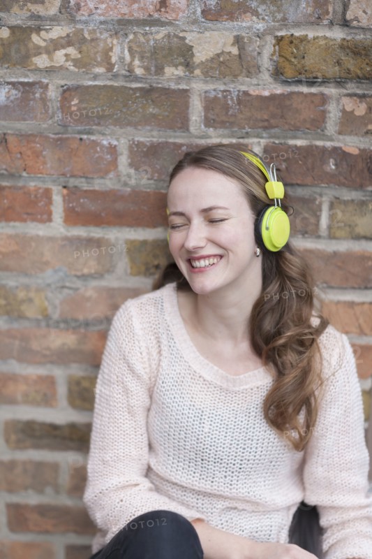 Mid-adult woman sitting and leaning against brick wall with closed eyes and headphones on