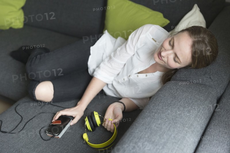 Mid-adult woman lying on sofa and holding smart phone and headphones