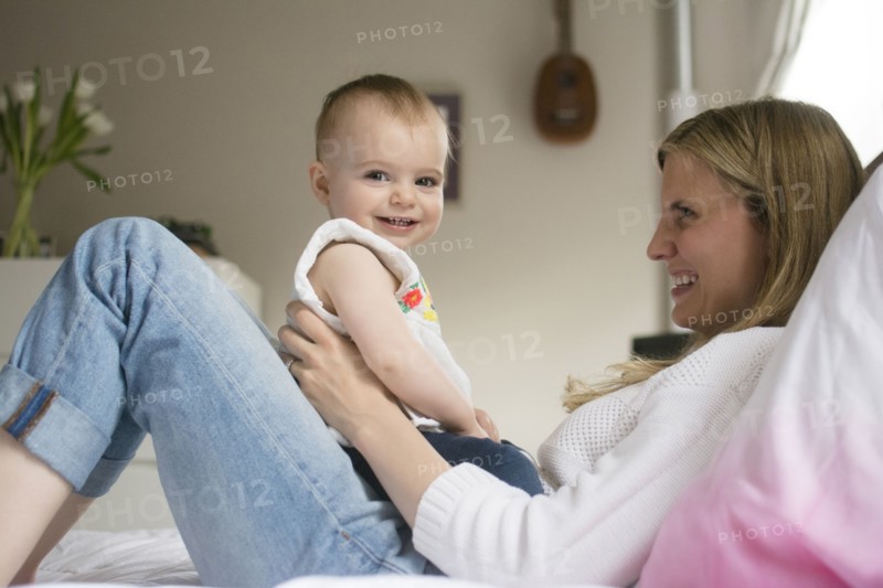 Mother and daughter (18-23 months) sitting on bed and smiling