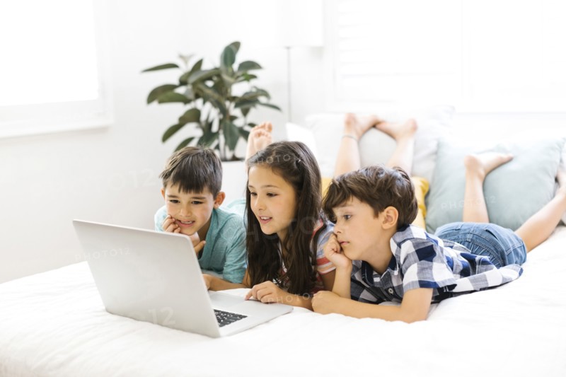 Siblings (10-11, 6-7, 8-9) lying in bed and using computer