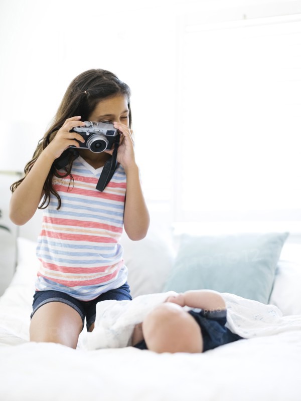 Girl (10-11)  taking photo of her small brother (12-17 months)  lying on bed