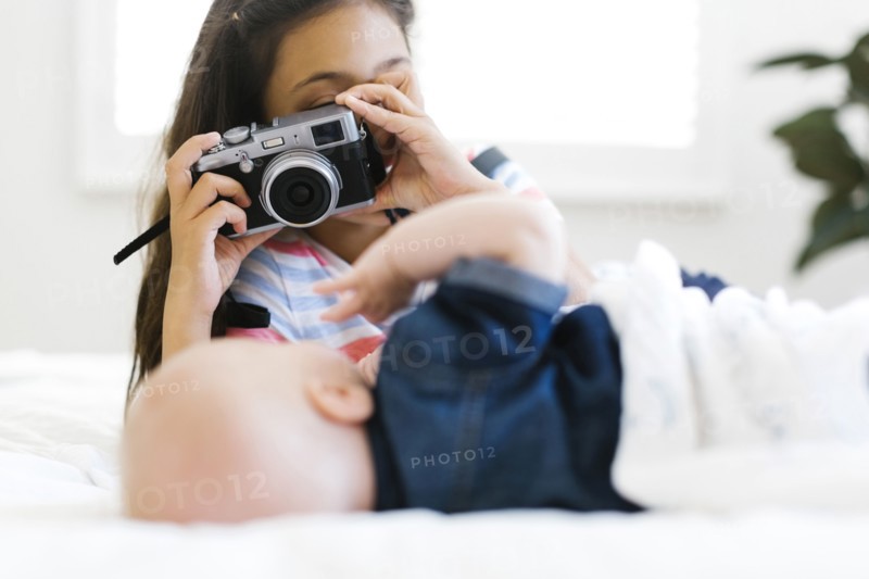 Girl(10-11) taking photo of her small brother (12-17 months) lying on bed