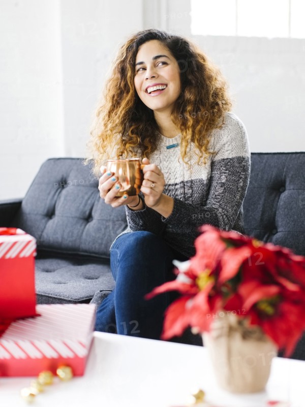 Happy woman on sofa, holding mug with Christmas gifts in foreground