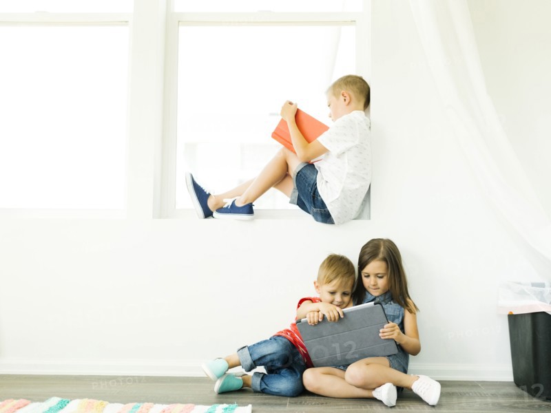 Boy (6-7) reading book and sister with brother (2-3, 6-7) using digital tablet