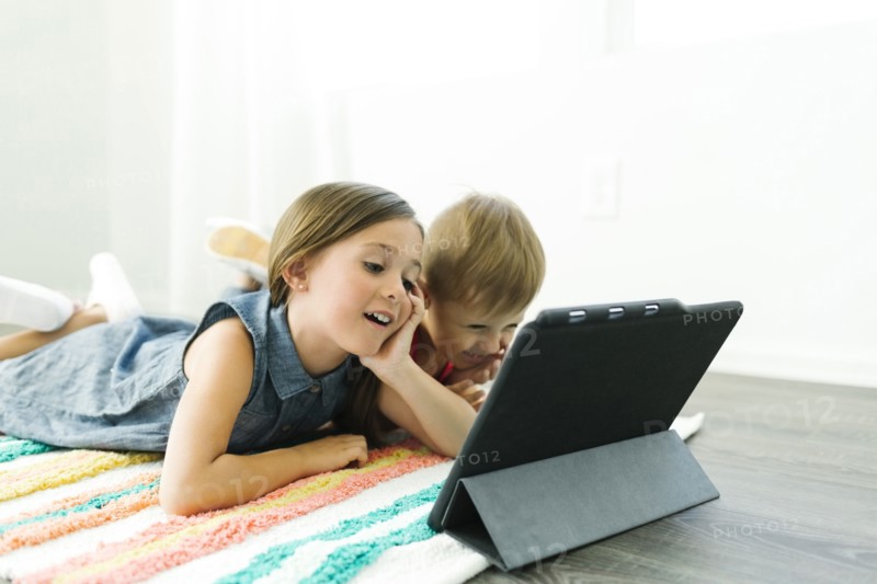 Siblings (2-3, 6-7) lying down on carpet and watching digital tablet
