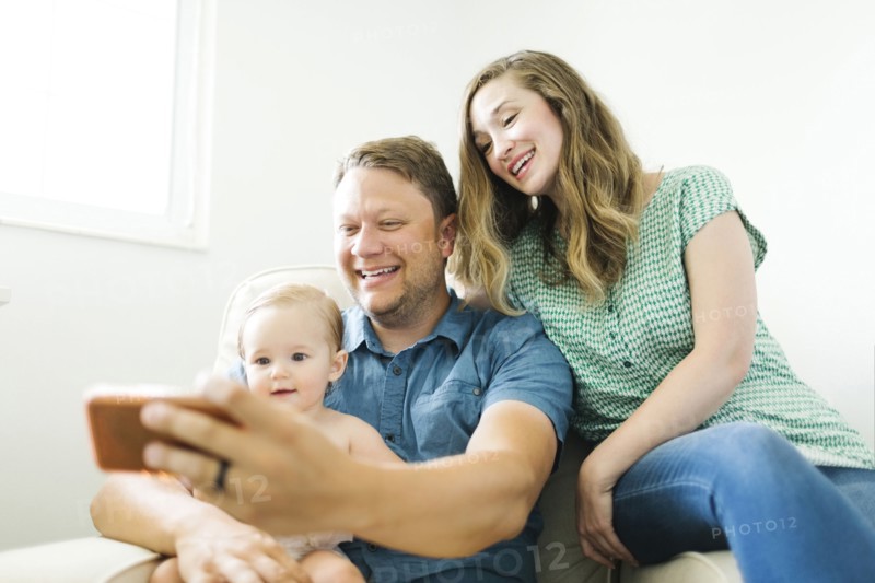 Mother and father with baby girl (12-17 months) sitting in living room and taking selfie