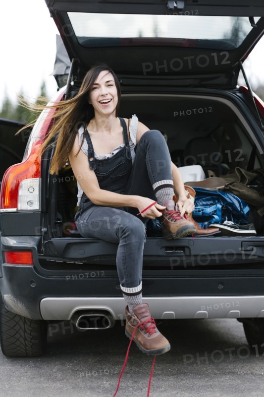 Smiling woman sitting in car trunk and tying hiking shoes