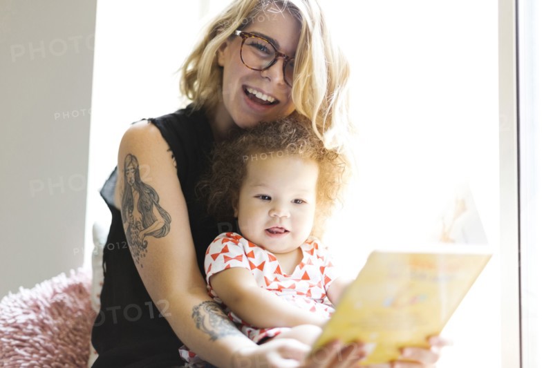 Mother sitting with daughter (12-17 months) on windowsill and reading book