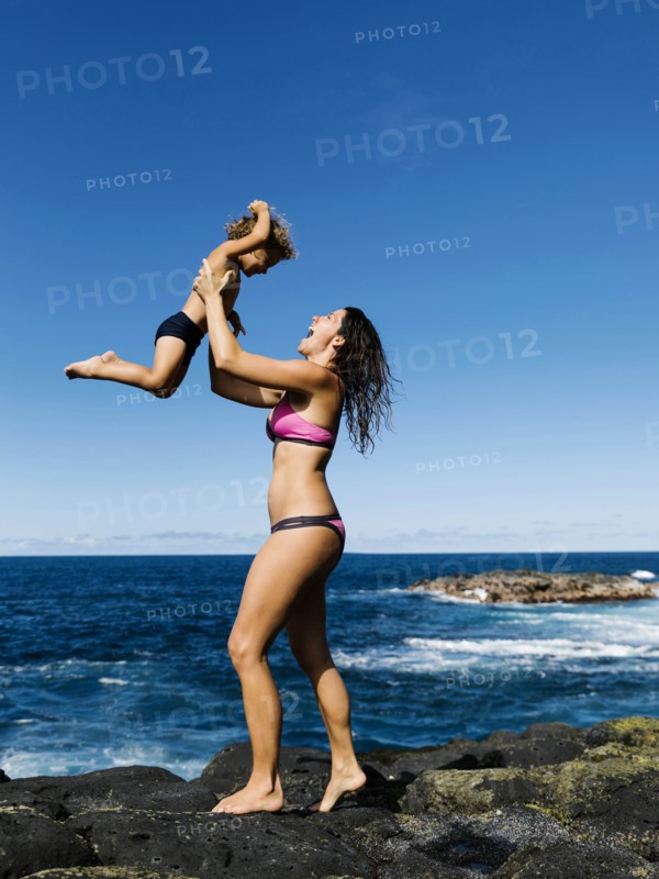 Mother lifting her son on rocks by sea