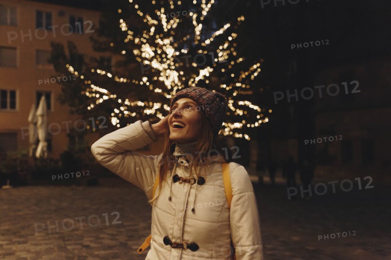 Smiling woman wearing coat by fairy lights in tree at night