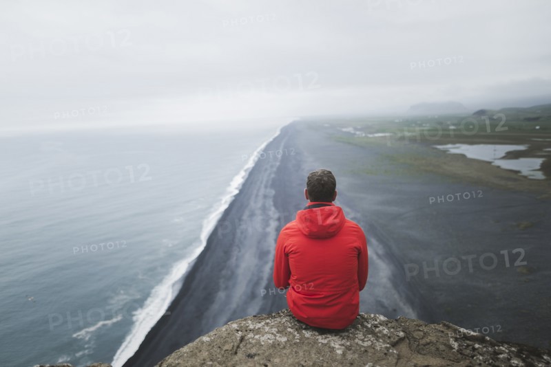 Man wearing red coat sitting above beach in Vik, Iceland