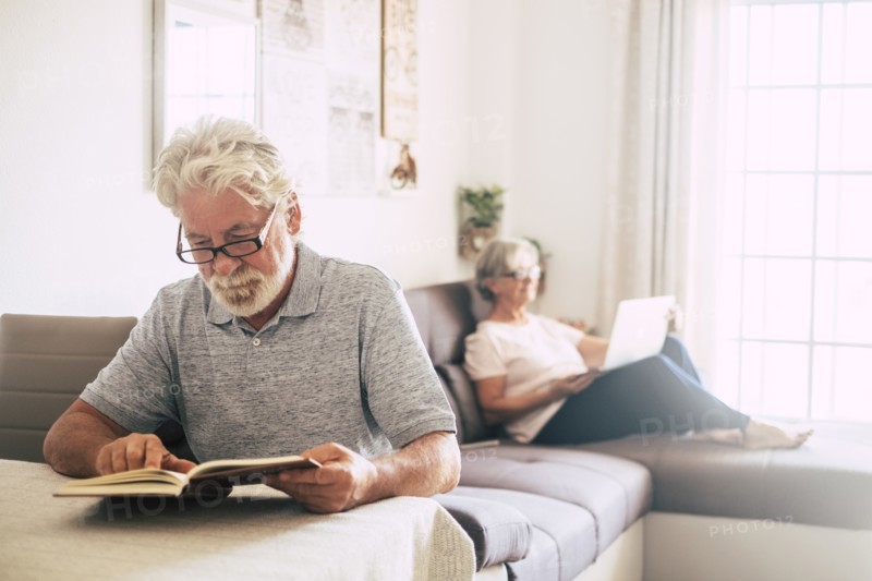 Senior man reading book as his wife uses laptop in living room