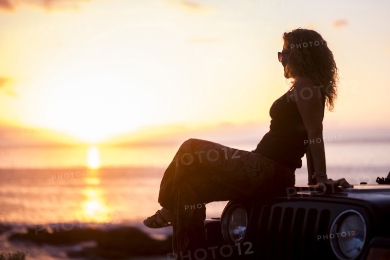 Woman sitting on car by beach at sunset