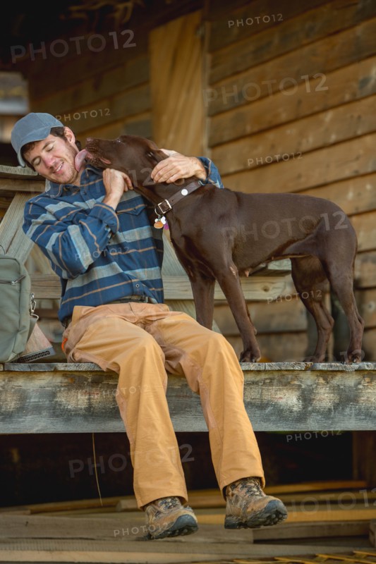 Chocolate Labrador licking man's face on porch of log cabin