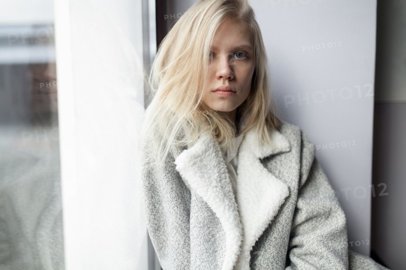young woman in wool coat leaning against wall by window