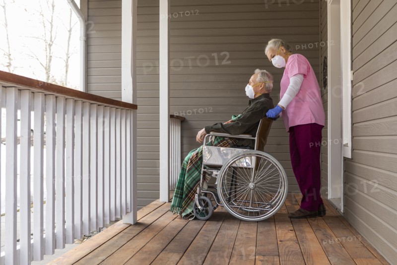 Woman and man in wheelchair wearing protective mask to prevent coronavirus transmission on porch