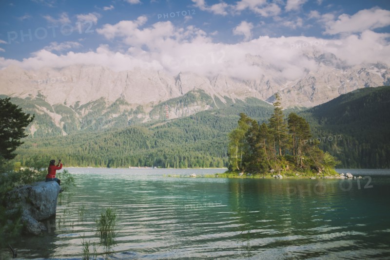 Germany, Bavaria, Eibsee, Young woman sitting on rock by Eibsee Lake in Bavarian Alps and photographing landscape with smart phone