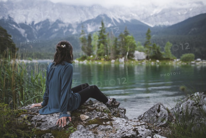 Germany, Bavaria, Eibsee, Young woman sitting on rock at shore of Eibsee lake in Bavarian Alps