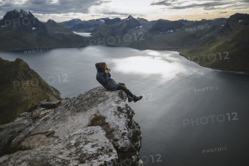 Norway, Senja, Man taking photo sitting on edge of steep cliff on top of mountain Segla