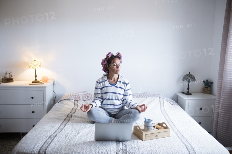 Woman with hair curlers meditating on bed in front of laptop