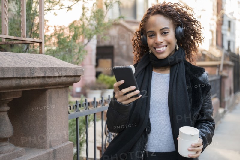 Smiling woman wearing headphones holding smart phone and cup