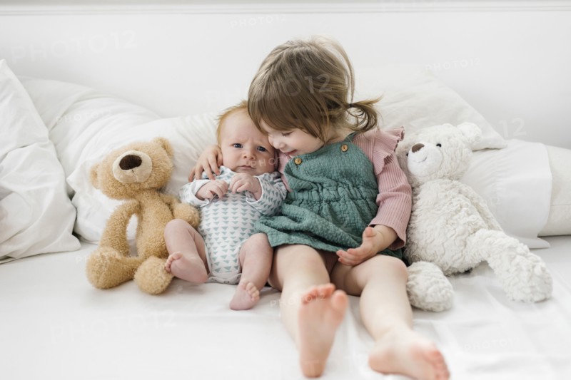 Siblings (2-5 months, 2-3) sitting on bed with teddy bears