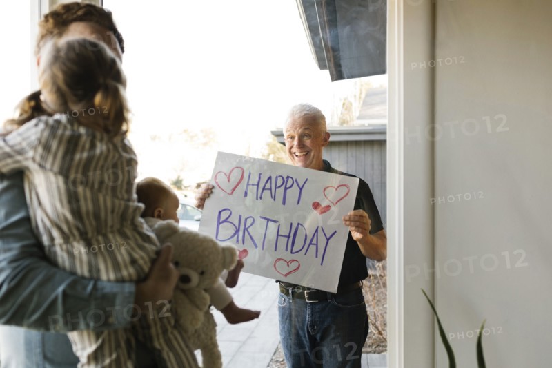Grandfather showing happy birthday message to family with grandchildren (2-3 months, 2-3) through window